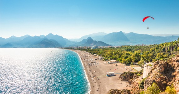 Dlouhá pobřežní pláž s průzračným mořem a horským horizontem zatímco nad krajinou letí paraglidista s červeným padákem. Slunečné středomořské pobřeží zachycuje dobrodružnou atmosféru dovolené v Turecku.