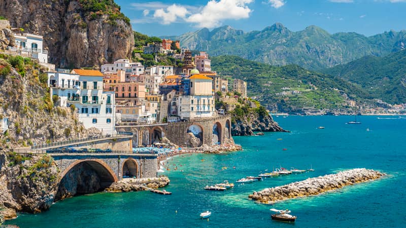 Barevné domy přilepené na skalnatém útesu nad tyrkysovým mořem a kamenný viadukt vedoucí podél pobřeží v oblasti Amalfi. Malebné středomořské panorama typické pro dovolenou v Itálii.